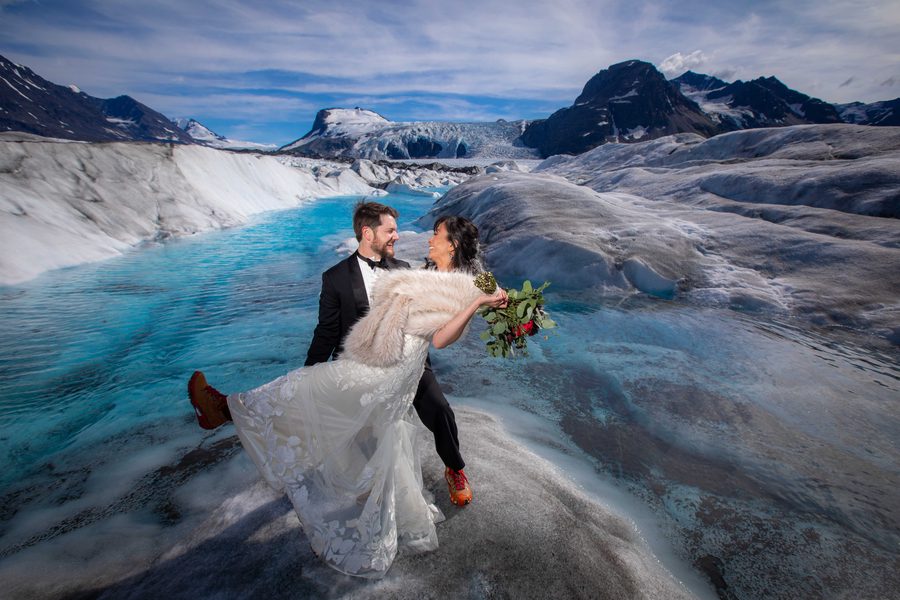A couple in wedding clothes laughing together on blue glacier ice.