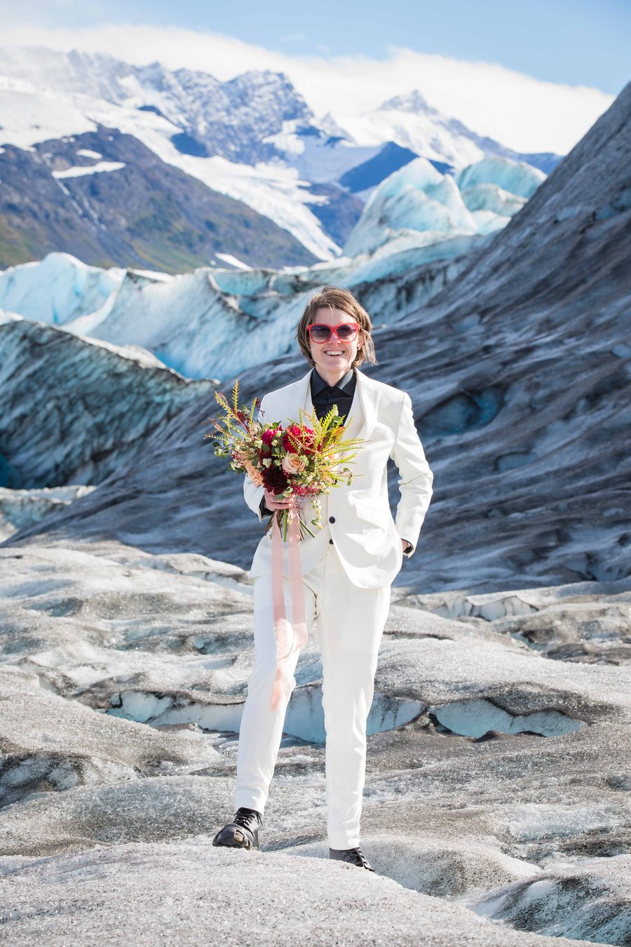 A couple standing together on glacier terrain with blue ice and mountains behind them.