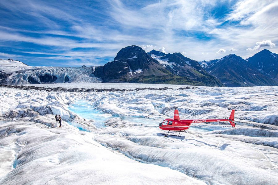 A red helicopter landed on a glacier beside blue ice and mountains.